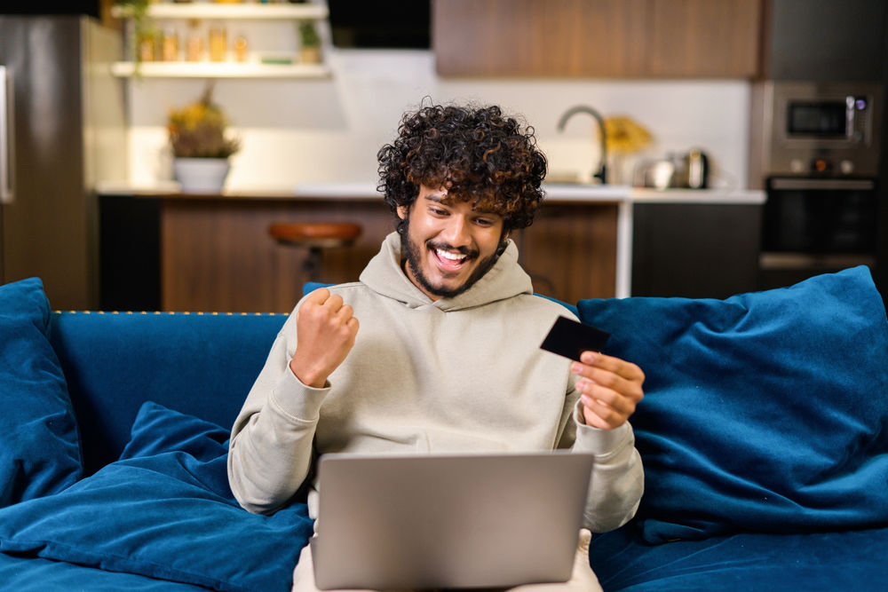 Smiling man holding banking card and laptop, making purchases or ordering online.