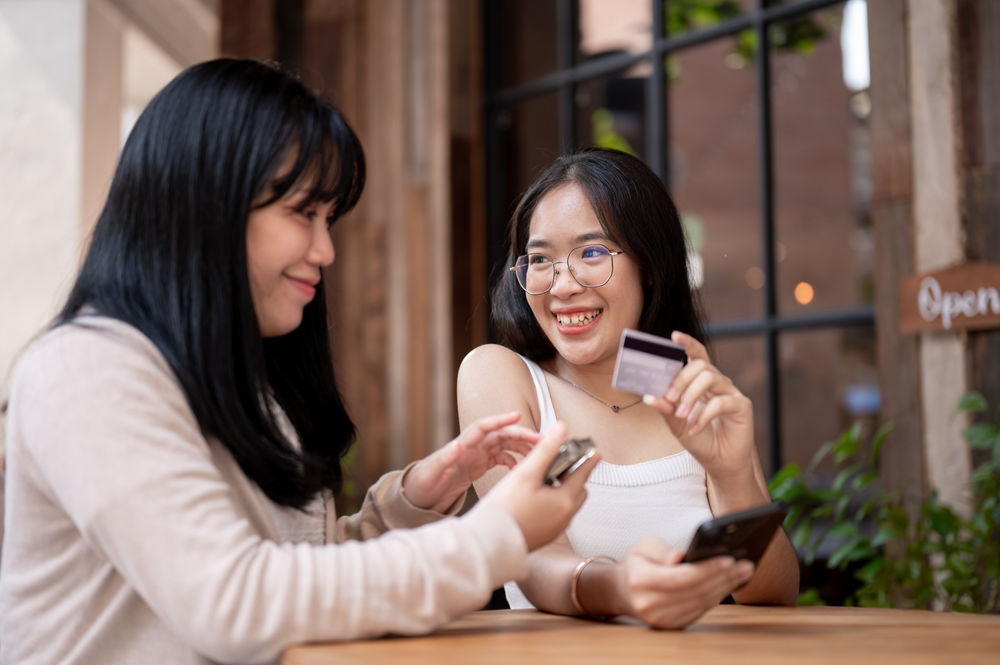 Two happy Asian female friends are talking after making an online hotel booking via a website with a credit card.